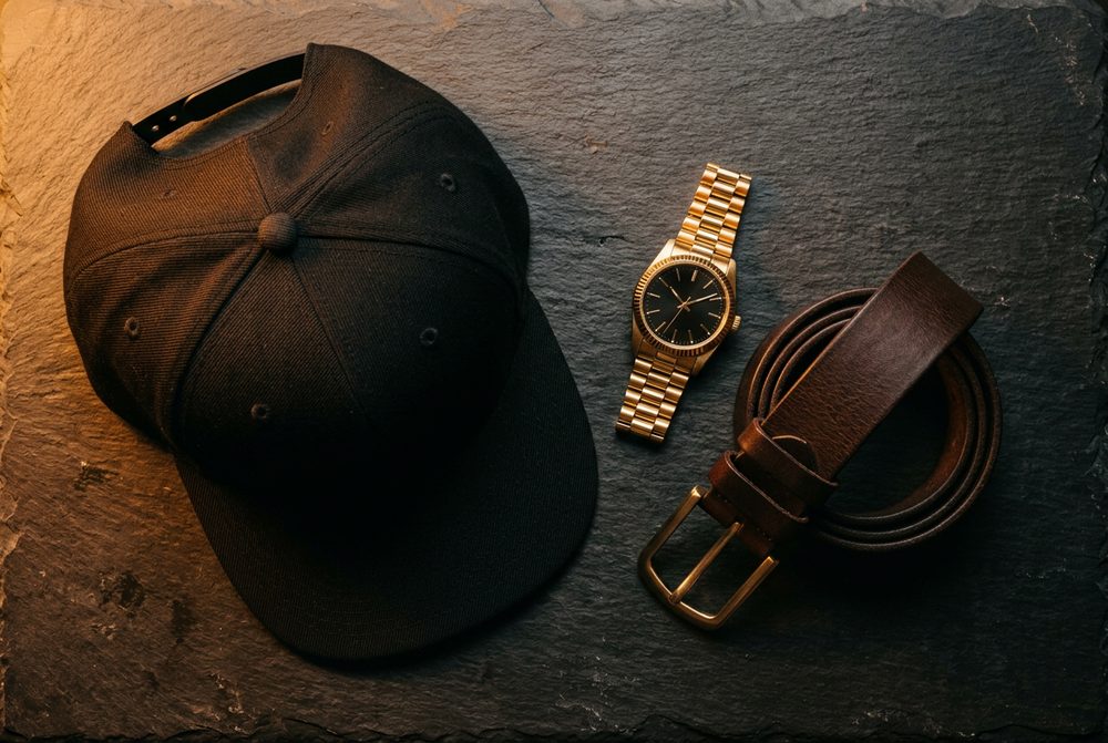 Gorra negra, reloj dorado y correa de cuero marrón con hebilla dorada, sobre una superficie de pizarra